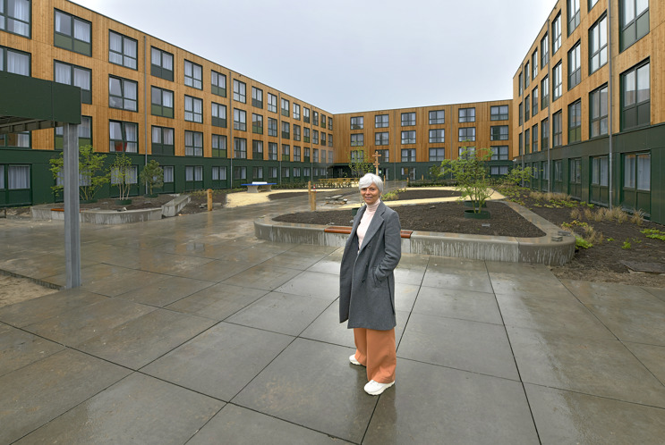 Caroline, grijze jas, bruine broek, poseert met handen in zakken voor het nieuwe appartementengebouw Bloemkwartier in Tilburg